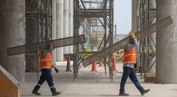 Dos trabajadores de construcción, debidamente vestidos, cargando estructuras metálicas en sus hombros, dentro de un ambiente de construcción